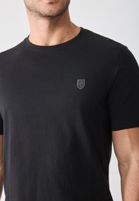 Black cotton t-shirt with a round neckline and short sleeves. Features a small gray shield logo on the left chest area. Soft texture.