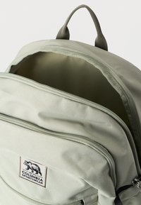 Ανοιχτό γκρι σακίδιο της Columbia Sportswear με μπροστινή τσέπη με φερμουάρ και επάνω χειρολαβή, μερικώς ανοιχτό κύριο διαμέρισμα που δείχνει το εσωτερικό ύφασμα επένδυσης.