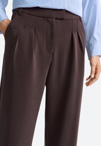 Pantaloni beige plissettati con una texture di tessuto liscia, dotati di tasche laterali e una vestibilità su misura. Abbinati a una camicia a righe blu e bianche.
