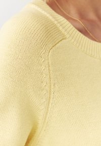Jasnożółty sweter z dzianiny z prążkowanym dekoltem i teksturowanym szyciem. Zbliżenie pokazuje gładki, miękki materiał i detale szwów.