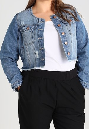 Femme portant une veste en jean courte bleue avec des boutons en cuivre, chemise blanche et pantalon noir taille haute, mains dans les poches.