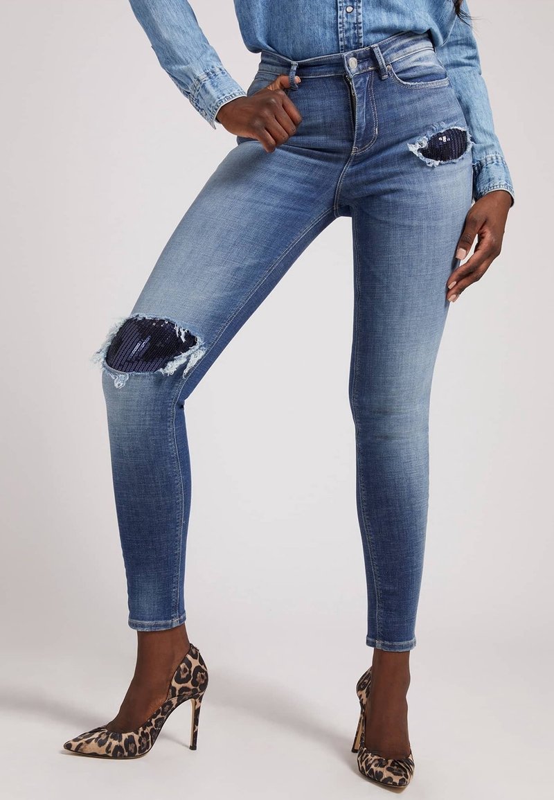 Jeans ajustados en denim azul de lavado medio, con una rodilla desgastada y lentejuelas azules marinas, bordes deshilachados, y combinados con tacones de estampado de leopardo.