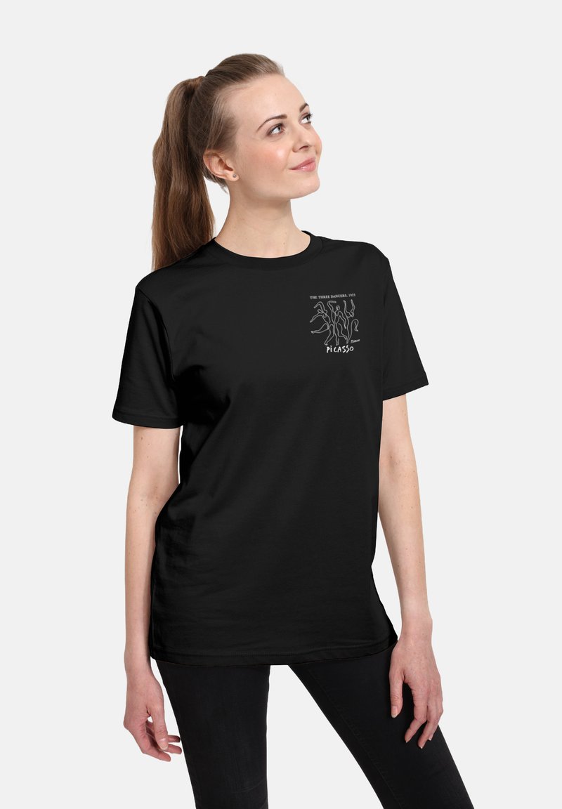 Mujer con coleta marrón que lleva una camiseta negra con un dibujo en línea blanca titulado "Las tres bailarinas, 1925" de Picasso.