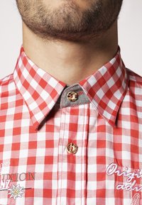 Camisa a cuadros rojos y blancos, con cuello abotonado, detalle bordado y botón de contraste con diseño de piel de leopardo.