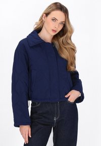 Kvinde med langt blont hår iført en marineblå quiltet jakke og mørke jeans, poserende med den ene hånd i lommen mod en hvid baggrund.