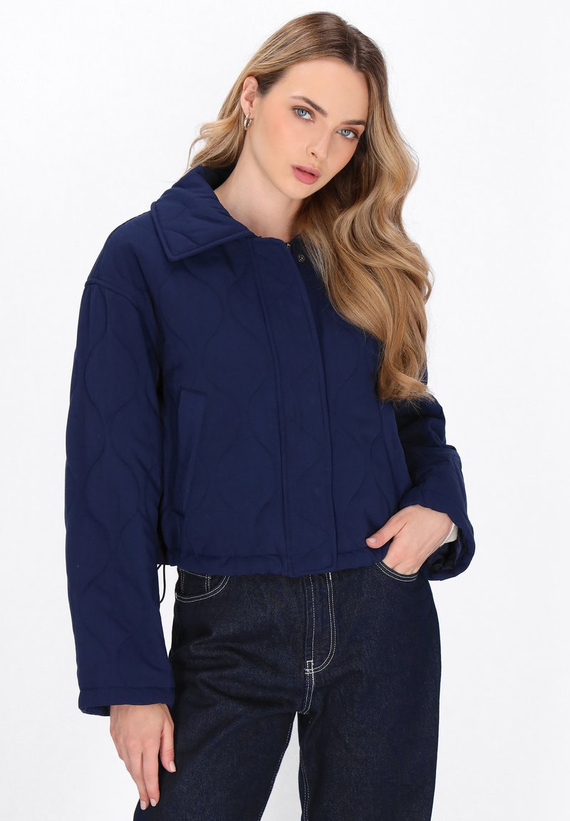 Kvinde med langt blont hår iført en marineblå quiltet jakke og mørke jeans, poserende med den ene hånd i lommen mod en hvid baggrund.