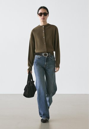 Brun cardigan med knapper, blå jeans med vide ben, sort bælte og solbriller. Modellen bærer en sort taske. Enkel, afslappet påklædning.