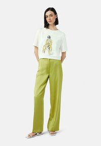 Weißes T-Shirt mit einer gedruckten Illustration, kombiniert mit hoch taillierten, weiten, limegrünen Satin-Hosen und weißen Riemchensandalen.