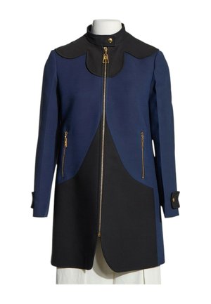 Abrigo largo azul y negro con cuello alto, cremallera dorada frontal, bolsillos con cremallera y puños con botones en un maniquí blanco.