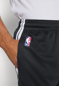 Shorts de basketball noirs en tissu maillé avec des rayures blanches sur les côtés et un patch du logo NBA sur la cuisse gauche. Taille élastique simple.