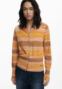 Stickad zip-up kofta i orange, gula och bruna ränder; har ribbade muddar och nederkant med broderad logotyp längst ner till vänster.