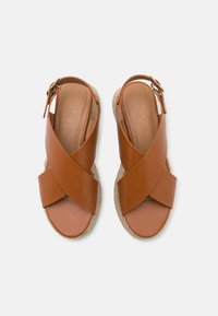 Paire de sandales en cuir beige avec brides croisées, fermetures à boucle réglables et semelles en espadrille tissée, sur fond blanc.