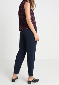 Marineblauwe broek gecombineerd met een mouwloze, donkere top met een rood hartpatroon. De outfit wordt aangevuld met zwarte schoenen met zilveren studs.