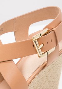Sandalia de cuña de cuero marrón con tiras cruzadas, detalle de hebilla dorada y suela de yute texturizada. Acabado suave con costuras mínimas visibles.