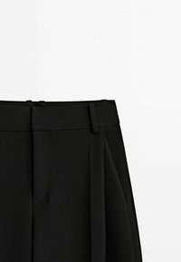 Pantalon noir taille haute sur mesure avec passants de ceinture et large ceinture latérale attachée, sur fond blanc uni.