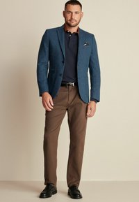 Hellblauer, strukturierter Wollblazer über einem marineblauen Polo, kombiniert mit braunen Hosen und schwarzen Schuhen. Verfügt über eine Brusttasche und Knöpfe.