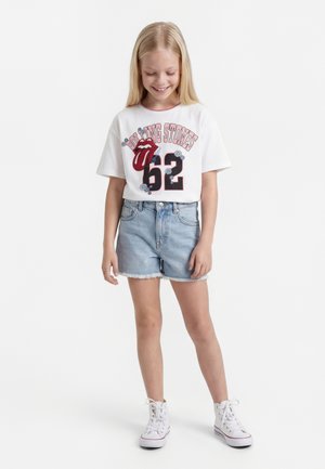 Νεαρό κορίτσι χαμογελά, φοράει λευκό T-shirt των Rolling Stones με τον αριθμό 62, ανοιχτόχρωμο τζιν σορτς και λευκά ψηλά αθλητικά, στέκεται μπροστά σε λευκό φόντο.