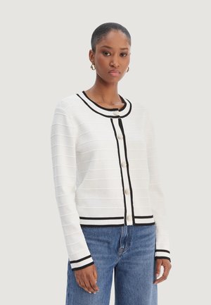 Kobieta ubrana w biały dziany kardigan z czarną lamówką i perłowymi guzikami, w połączeniu z niebieskimi dżinsami, stojąca na jednolitym tle.