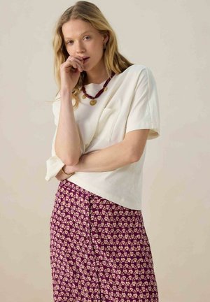 Witte top met korte mouwen en een zak, gecombineerd met een paarse rok met een bloemenpatroon. Een ketting met gouden accenten maakt de look compleet.