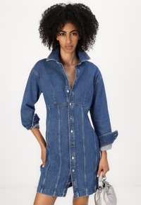 Kjole i mørkeblå denim med knappelukking, stor krage og oppbrettede ermer. Har kontrasterende sømmer og en formsydd midje.