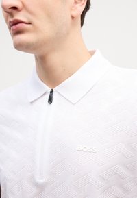 Bílá polo košile s texturovaným geometrickým vzorem, krátkým límečkem, polovičním zipem a diskrétním logem "BOSS" na hrudi.