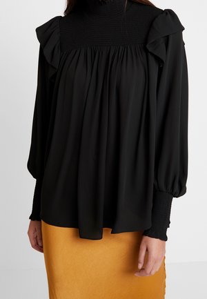 Frau trägt eine schwarze Bluse mit langen Ärmeln, gerafften Schultern und gerafften Bündchen, kombiniert mit einem senffarbenen Rock.