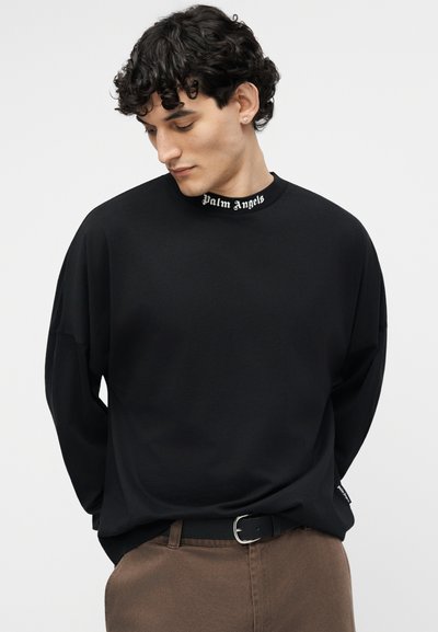 Giovane uomo con capelli ricci scuri che indossa una felpa nera con la scritta "Palm Angels" sul colletto e pantaloni marroni con cintura nera.