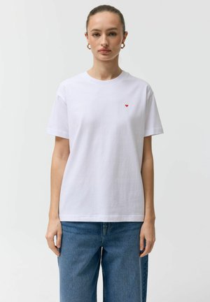 Kobieta z zawiązanymi włosami nosi prostą białą koszulkę z małym czerwonym sercem na piersi i niebieskie dżinsy, stojąc na jednolitym tle.