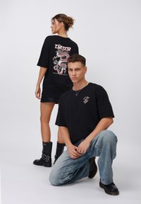Fekete, over-size t-shirt, amelynek hátulján egy piros sárkány dizájn található, elöl pedig egy kisebb grafika látható. A modell világoskék farmernadrággal viseli.