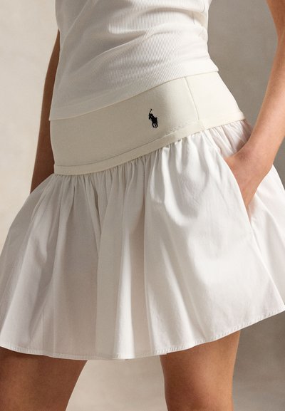 Kvinne iført et hvitt skjørt med vidde og rynket stoff, med en beige linning og en liten svart logo på hoften, hånden i lommen.