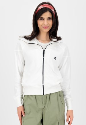 Young woman with dark hair and red polka dot headband wearing white zip-up hoodie and green cargo pants, standing against a plain background.