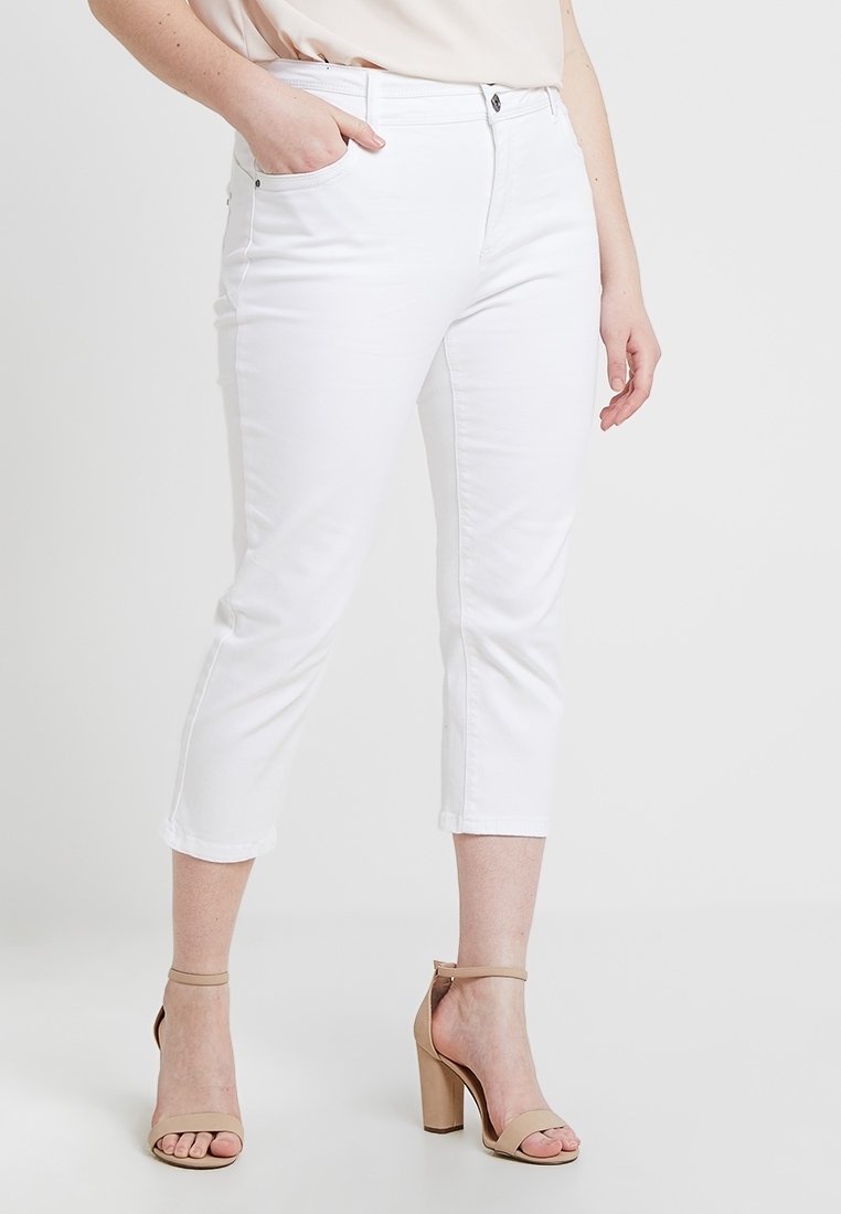 Femme portant un pantalon court blanc et des sandales à talons beiges, debout avec une main dans la poche, sur un fond uni.