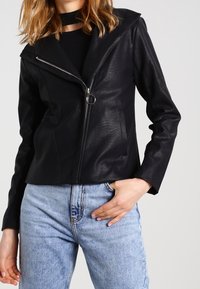 Chaqueta de piel negra con capucha puntiaguda, cremallera asimétrica y bolsillos frontales. Combinada con jeans de color azul claro que muestran un estilo ajustado.