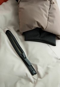 Veste rembourrée avec un extérieur beige, une fermeture éclair noire et des poignets en élastique sombres. Le tissu semble lisse avec une finition mate.