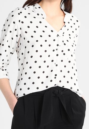 Valkoinen napillinen pusero, jossa mustia pilkkuja, taitetut 3/4-hihat, etutasku, yhdistetty korkea vyötäröisiin mustiin housuihin, jotka on sidottu vyötäröltä.