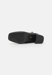 Chaussure noire avec un dessus en cuir lisse, bout carré, talon bas carré, et accent décoratif en métal sur le côté. Semelle extérieure en caoutchouc texturé.