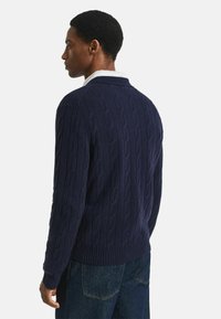 Man die naar links kijkt, gekleed in een marineblauwe kabeltrui over een wit shirt en donkere spijkerbroek, tegen een plain witte achtergrond.