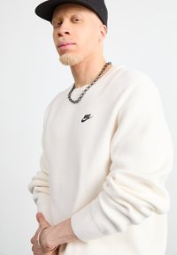 Valkoinen villapaita, jossa on ribbikujat, ja siinä on pieni musta Nike-logo. Korostettu paksulla mustalla ketjusolki-kaulaketjulla. Mallilla on päässään musta lippalakki.