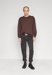 Braunes Langarmshirt, graue Sweatpants mit elastischen Bündchen und schwarze Sneakers. Das Outfit hat eine Kordelzugdetails an der Taille.