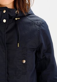 Chaqueta azul marino hecha de una tela ligera, con capucha, detalles en hardware dorado, un bolsillo con botón y cordón ajustable.