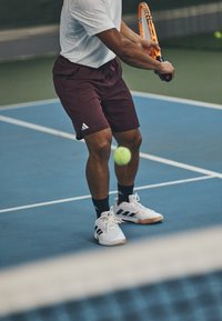 Joueur de tennis en chemise blanche et shorts bordeaux, tenant une raquette orange. Il porte des chaussettes noires et des chaussures blanches avec des rayures bleu marine.