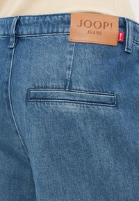 Jeans aus Denim in mittlerem Blau mit einem Lederlabel, das "JOOP! JEANS" liest. Verfügt über eine Gesäßtasche und eine leicht ausgewaschene Textur.
