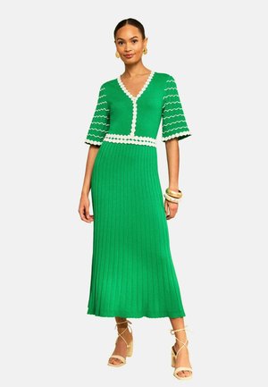 Frau trägt ein grünes Midikleid mit weißem Besatz, wellenförmigen Ärmelmustern, kombiniert mit beigen Sandaletten mit Absatz sowie goldenen Armbändern und Ohrringen.