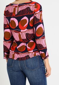 Haut multicolore avec des motifs abstraits en rose, rouge et marron sur un fond marine. Comprend des manches trois-quarts et une ouverture clé à l'arrière.