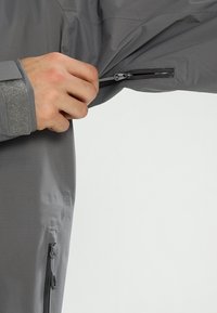 Main fermant une petite poche zippée sur la manche grise d'une veste, montrant un poignet avec une sangle en velcro et une autre poche en dessous.