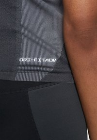 Tmavě šedý sportovní top bez rukávů s ribkovanou strukturou. Logo "DRI-FIT ADV" v bílé barvě na boku, spárovaný s černými těsnými legínami.