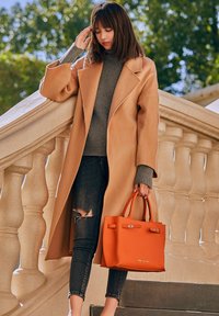 Cappotto cammello, maglione grigio a costine, jeans scuri con strappi e una borsa in pelle arancione con manici. Lo sfondo presenta una scalinata in pietra.