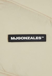 Chaqueta de plumas beige con una superficie texturizada, que cuenta con una etiqueta de goma negra que dice "MJGONZALES", exhibida de manera destacada en la parte frontal.