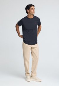 T-shirt di cotone blu navy con maniche corte, scollo rotondo e piccolo logo rosso, abbinata a pantaloni beige chiaro e scarpe da ginnastica color crema. Tessuto liscio.