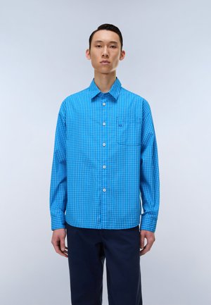 Mand stående iført en blå ternet langærmet skjorte med knapper og mørke bukser mod en ensartet lys baggrund.